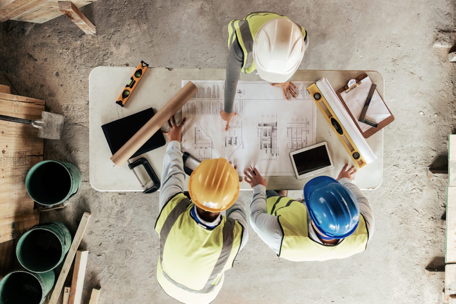 Overhead view of three construction workers in hard hats reviewing architectural blueprints on a table at an active construction site with measuring tools, a tablet, and rolled plans.